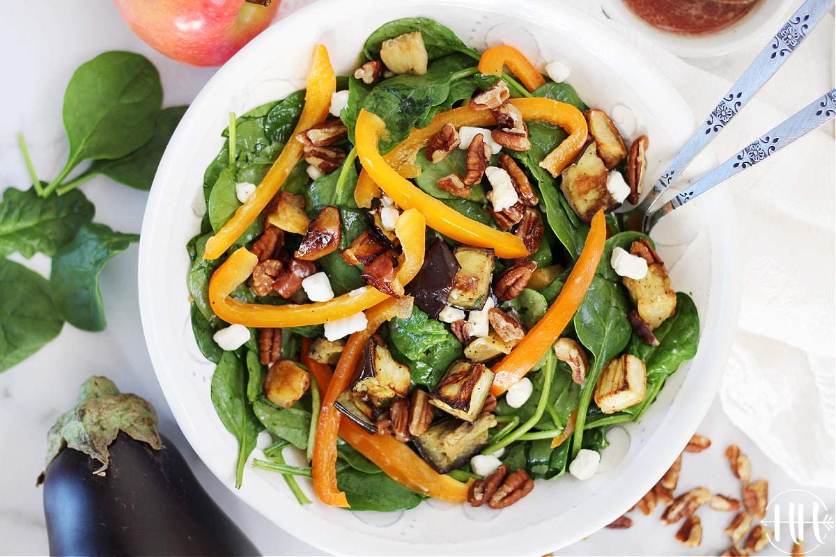 Tossed clean eating spinach salad with vegetarian toppings in a white bowl and a beautiful eggplant in the background.