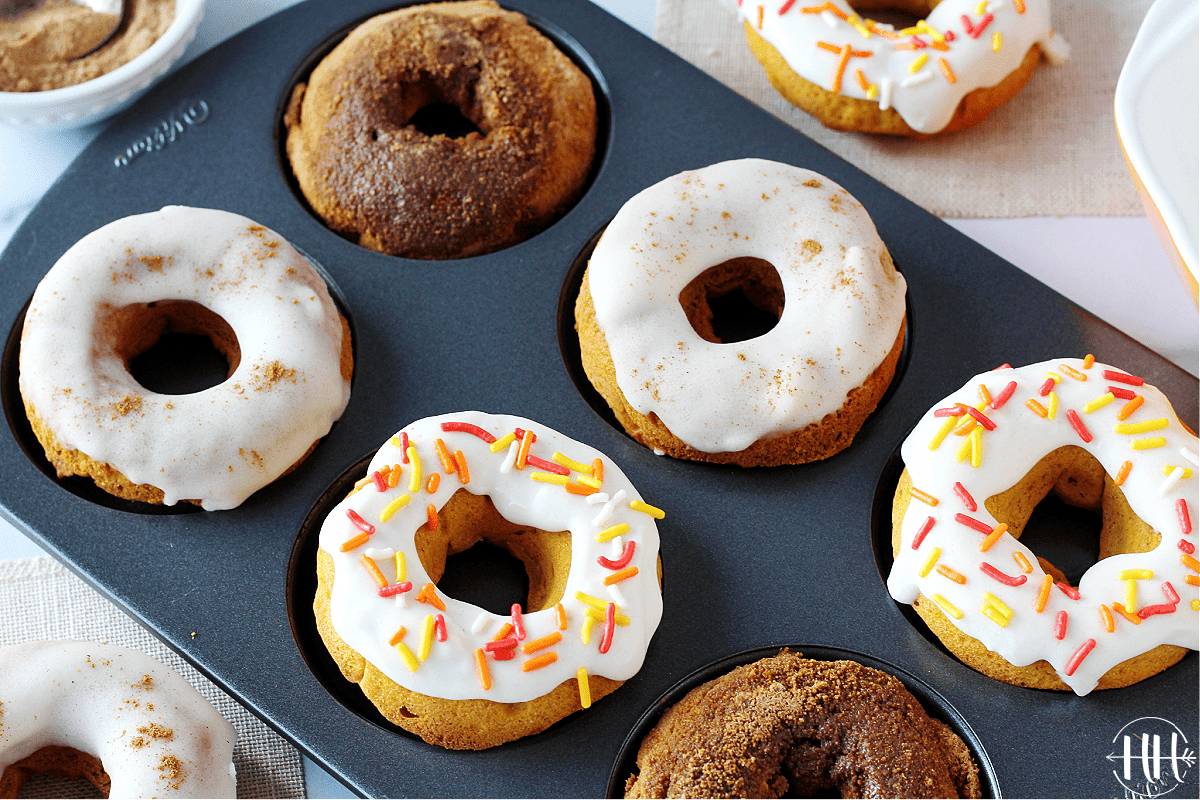 Six baked pumpkin donuts in a donut pan with different toppings and sprinkles.