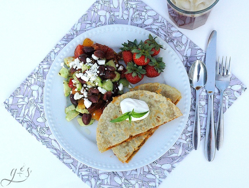 Over head shot of halved sloppy joe quesadillas on a white plate with a Mediterranean salad and fresh strawberries on the side.