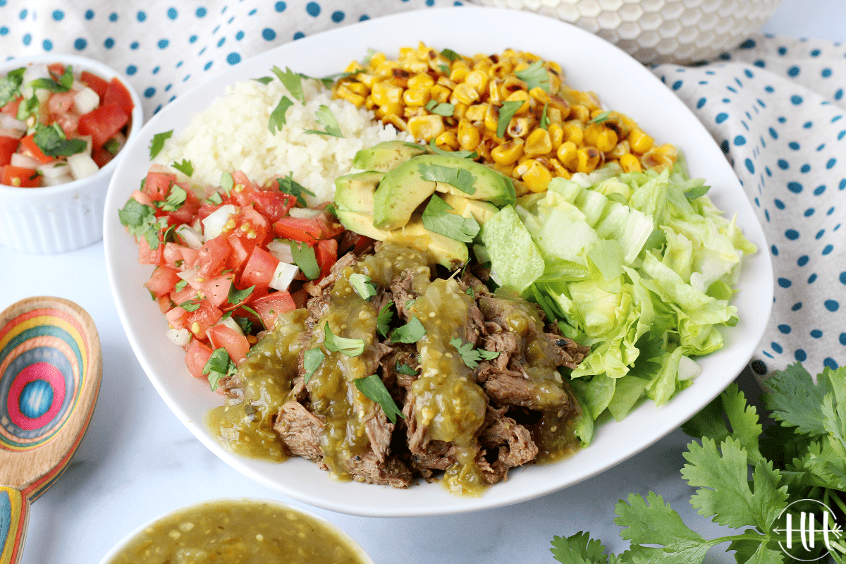 A beautiful bowl of riced cauliflower, pico de gallo, fire roasted corn, lettuce, avocado, and Mexican shredded beef surrounded by fresh cilantro and bowls of ingredients.