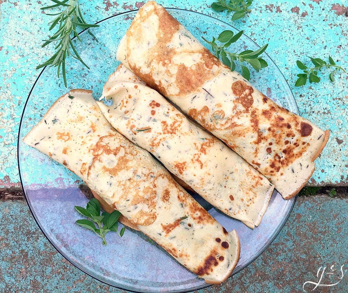 Three thin and savory crepes on a plate with garden herbs. 