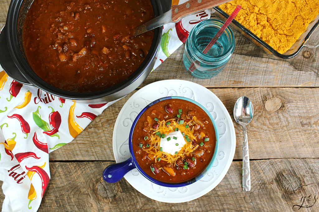 Birds eye view of a large pot of fall chili, pumpkin cornbread, and a bowl of warm chili topped with shredded cheese sour cream, and green onions. 