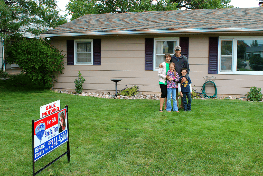 Family of four standing in front of their first home with a for sale sign in front of it. The home is tan with white trim and eggplant colored shutters. 