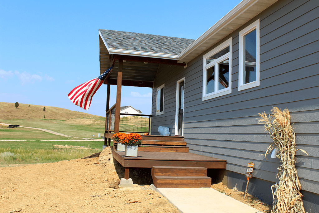 New home with grey siding and white trim. An American flag flying in the wind on the front porch. It's fall too with orange mums in rusted tin planters and a stack of cornstalks leaning against the home.