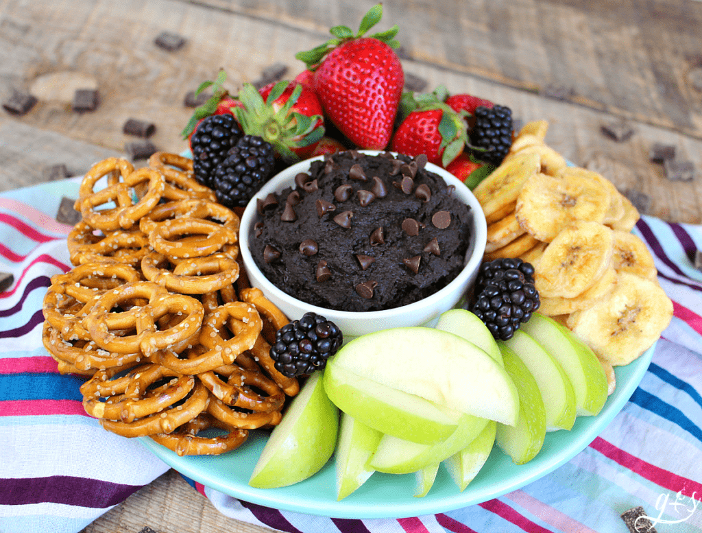 Dark chocolate dessert hummus in a bowl topped with chocolate chips surrounded by fruit and pretzels. 