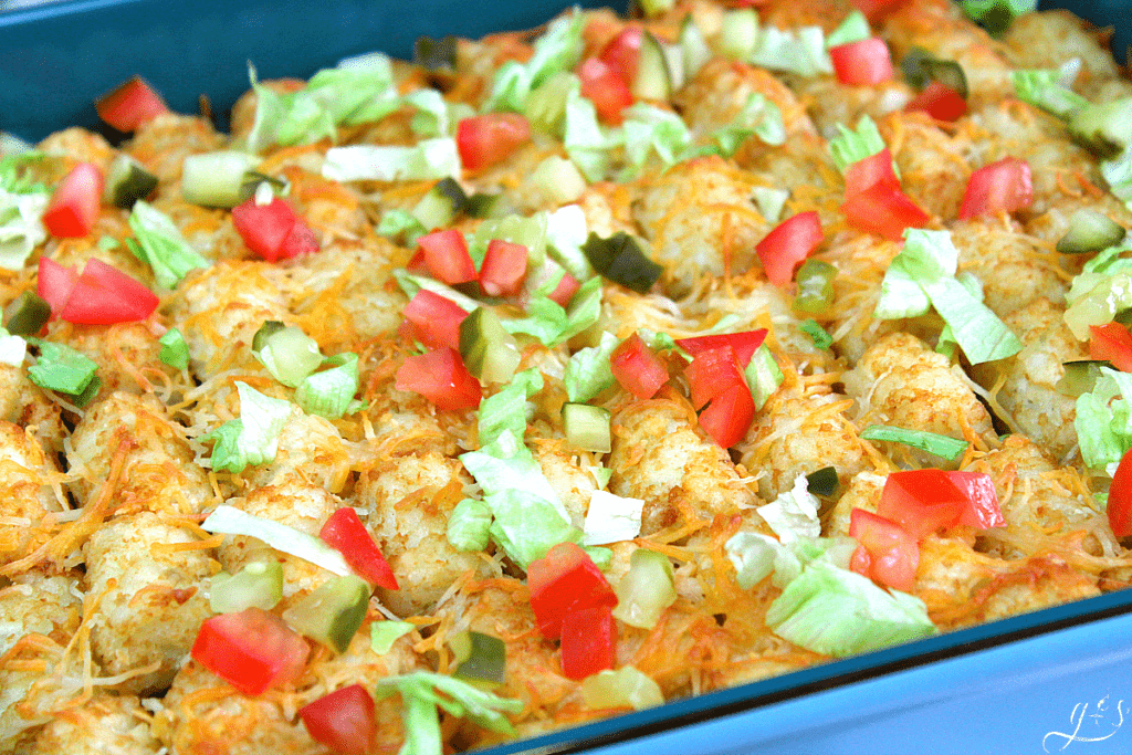 A pan of Tater Tot Topped Cheeseburger Casserole with pickles, tomatoes, and lettuce.