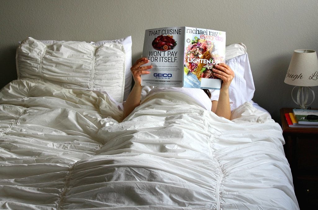 Girl lying in a white linen covered bed reading a Rachael Ray Everyday magazine. 