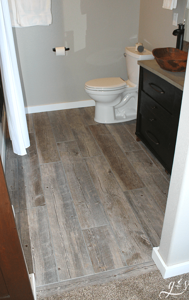 DIY How to Lay Floor Tile Planks | Our master suite bathroom floor is rustic and fun with the light tile that looks like wood and dark gray grout. These porcelain tile ideas and farmhouse designs are simple and beautiful. This tutorial to install your own large plank wood-look tiles on a budget will make you feel like a professional! I love the contrast of the grey-brown planks, white trim, dark vanity, and copper vessel sink. #farmhouse #tile #decor 