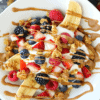 A breakfast banana split on a white plate surrounded by granola and fresh berries.