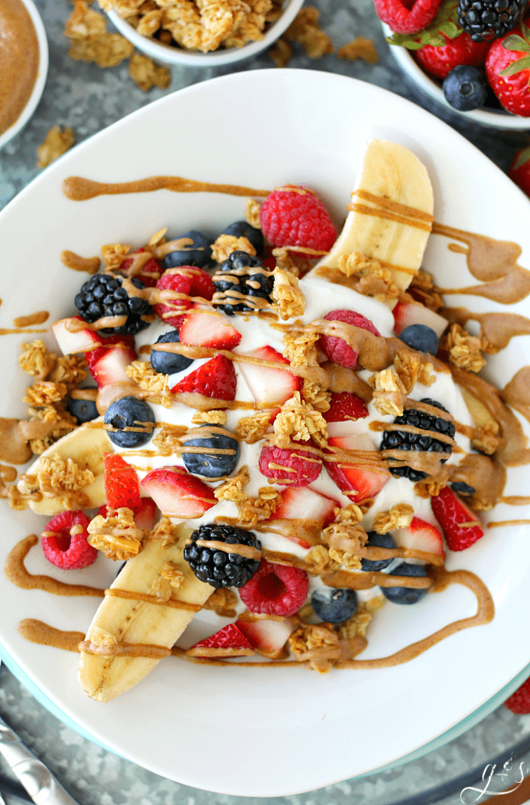 A breakfast banana split on a white plate surrounded by granola and fresh berries.