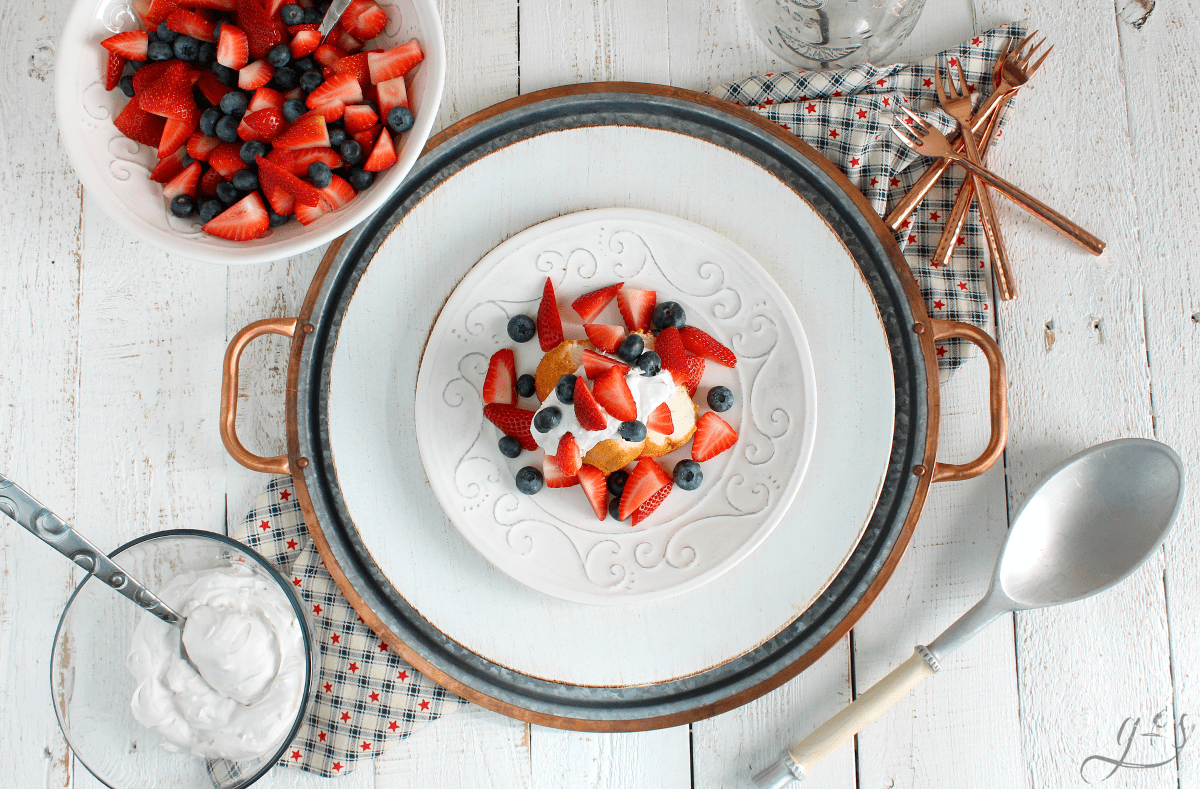 Patriotic Shortcake on a white cake with a bowl of berries, coconut whipped cream and various red, white, and blue props. 