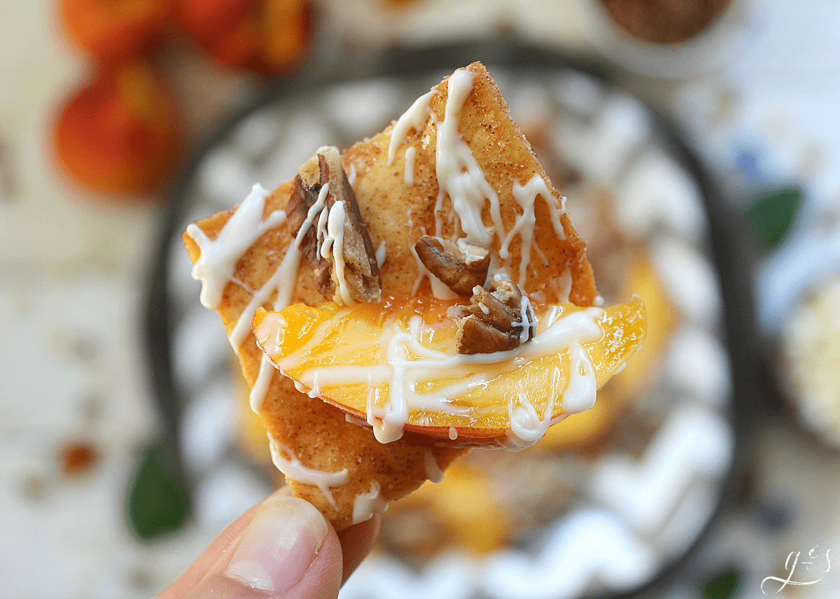 A cinnamon pita chip topped with melted white chocolate, a sliced peach, and chopped pecans. 