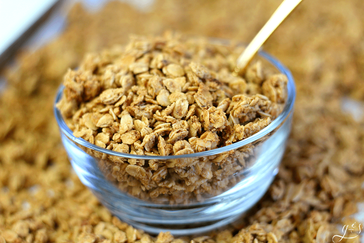 Clean glass bowl of French Toast Granola with a gold spoon in it atop a pan of the granola. 