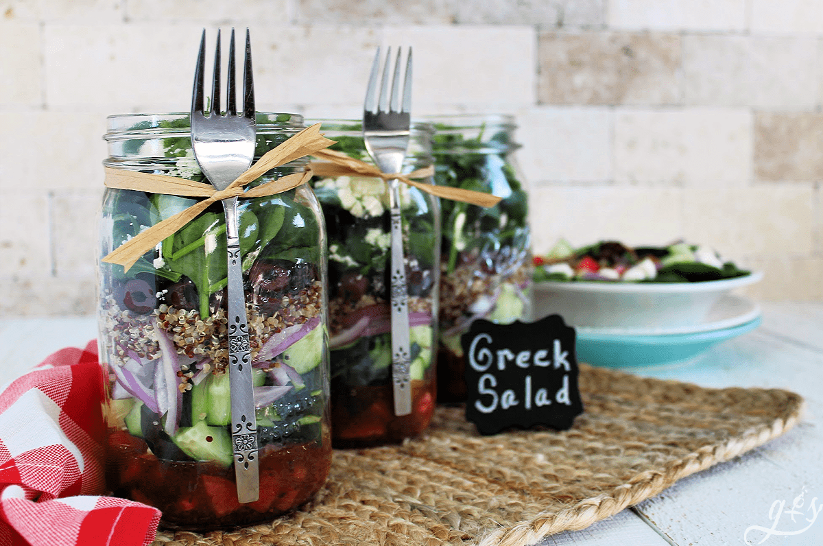 Three wide mouth mason jars filled with Greek Salad ingredients and a fork tied on each jar with raffia. 