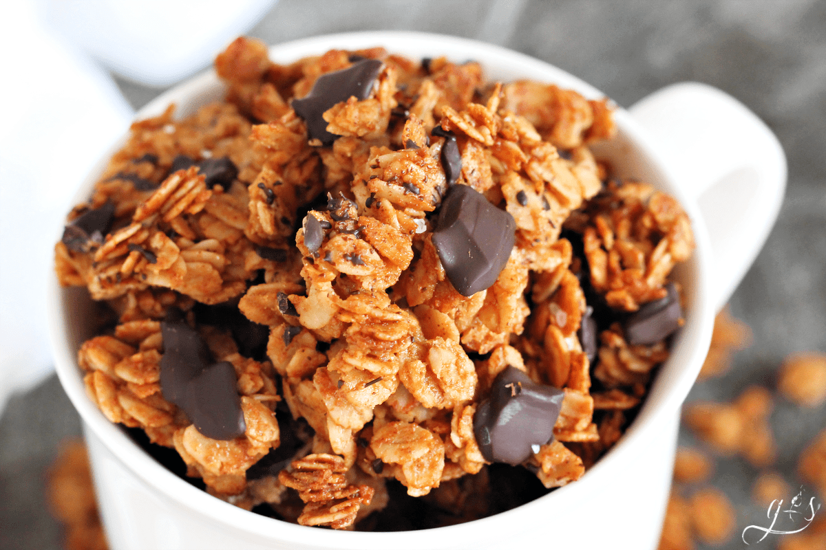 Close up of white mug full of almond butter granola with dark chocolate chunks.