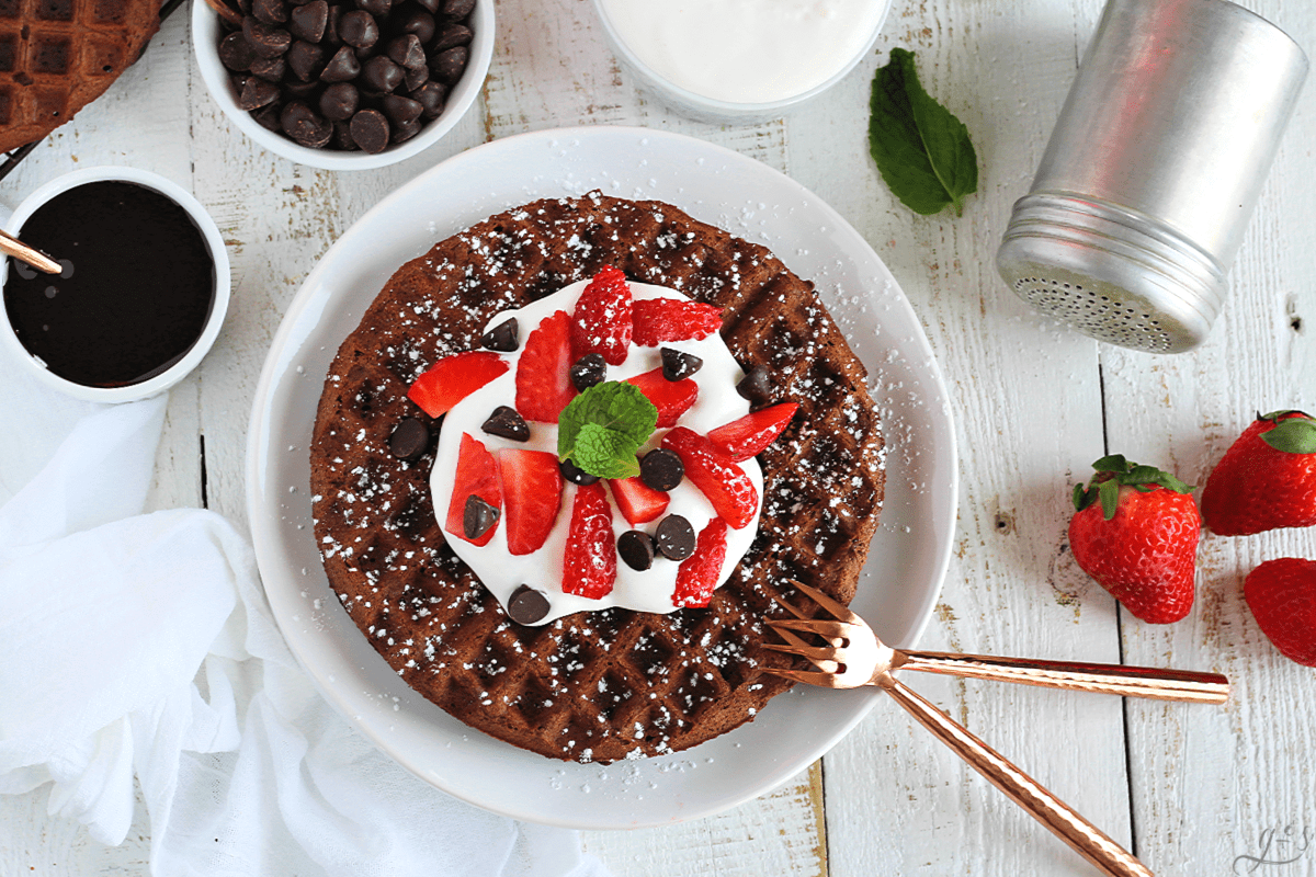 Overhead shot of gluten free chocolate waffles topped with whipped cream and fruit. Small bowls of chocolate sauce, chocolate chips, whipped cream, and a shaker with powdered sugar beside the plate.