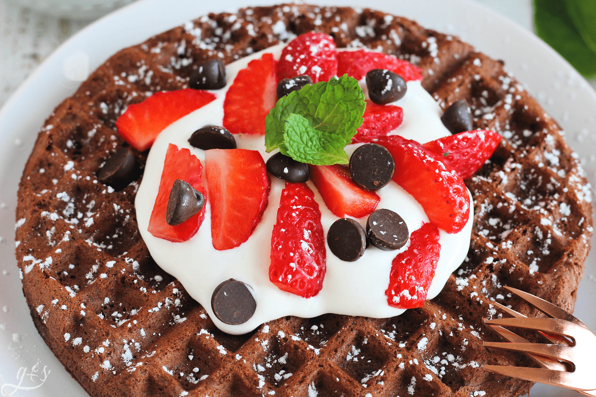 Up close photo of a homemade chocolate waffle topped with whipped cream, fresh strawberries, chocolate chips, and fresh mint. 
