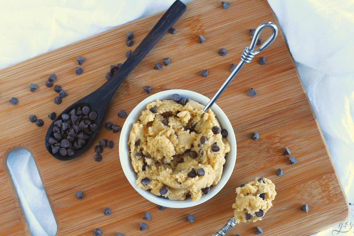 Overhead photograph of edible chocolate chip cookie dough in a white bowl atop a wooden cutting board.