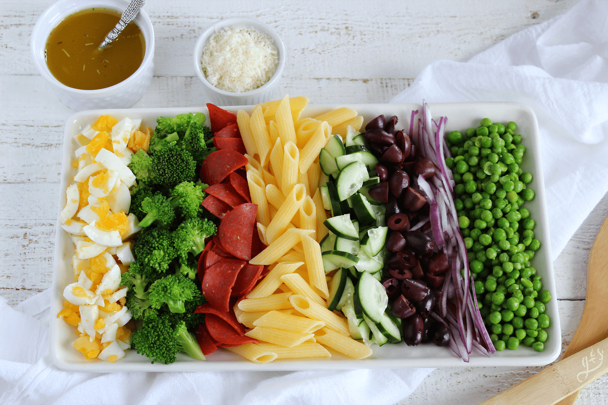 All the ingredients for a pasta salad including rows of veggies, olives, and pasta arranged neatly on a plate with a bowl of dressing and of Parmesan. 