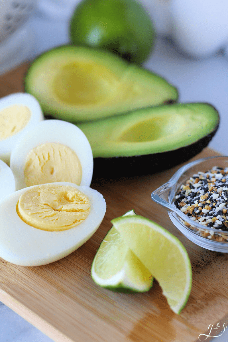 Close up photo of lime slices, hard boiled eggs, sliced avocado, and EBTB seasoning on a cutting board for an avocado egg salad.