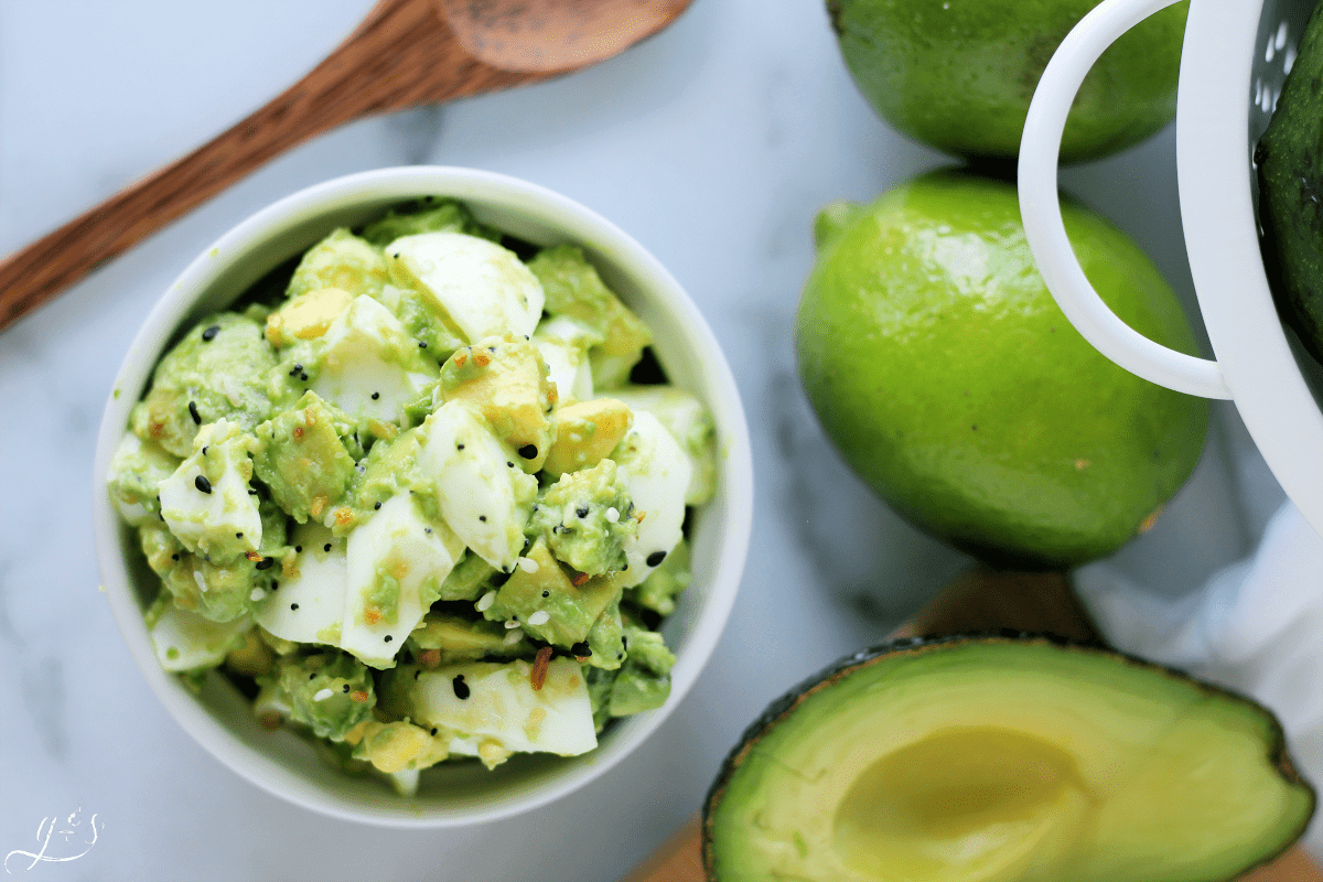 Overhead photo of a small white bowl filled with 4 Ingredient Avocado Egg Salad styled with a wooden spoon, lime and avocado half.