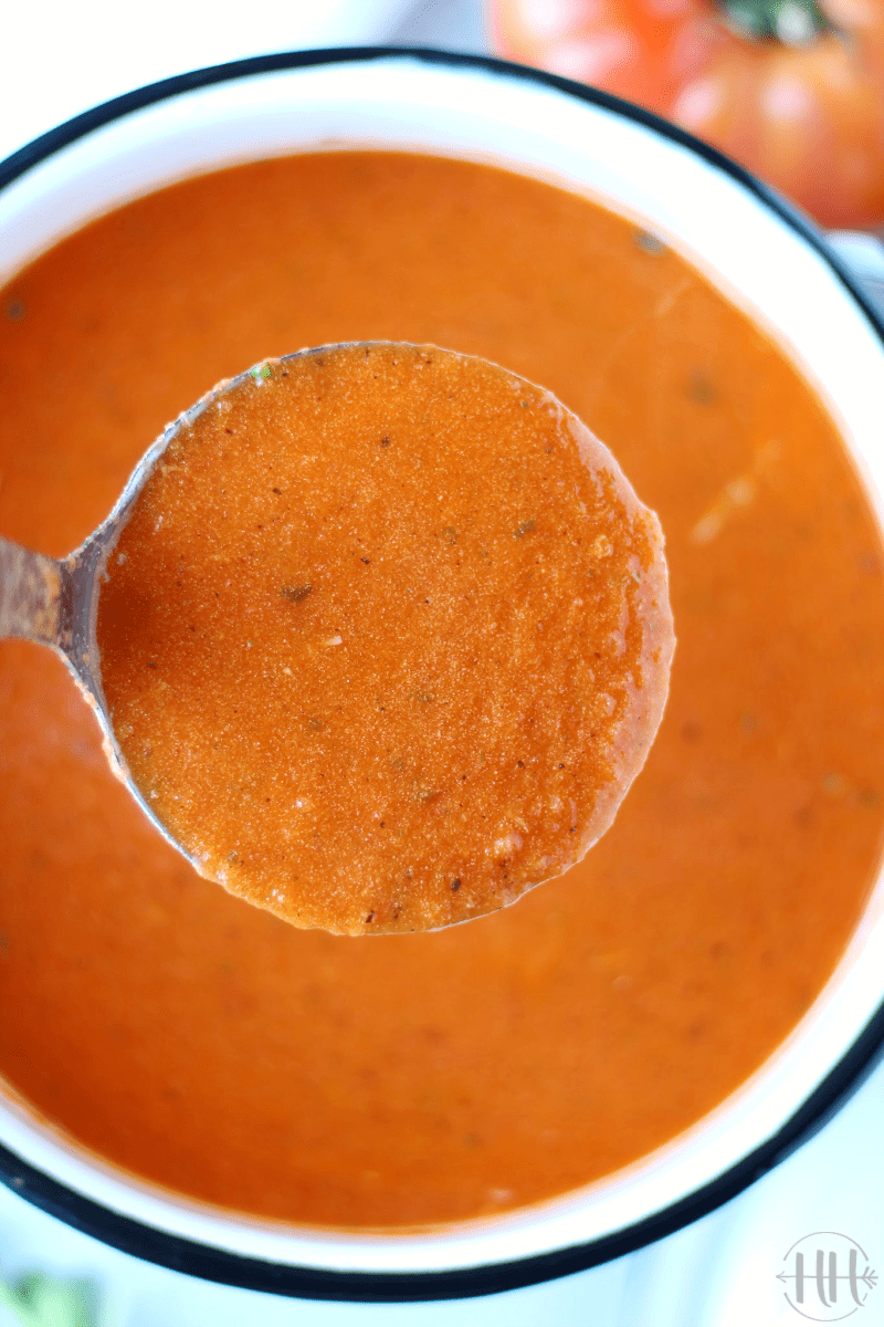 Overhead photo of a metal ladle of creamy homemade tomato soup in a white pot.