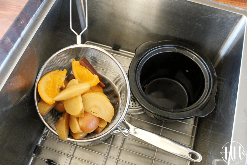 Cooked apples and spices in a metal strainer over a pot of apple cider in a metal sink. 