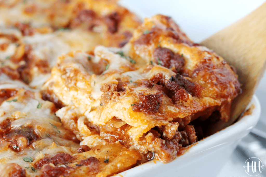 A wooden spoon scooping out a piece of homemade lasagna out of a white pan.