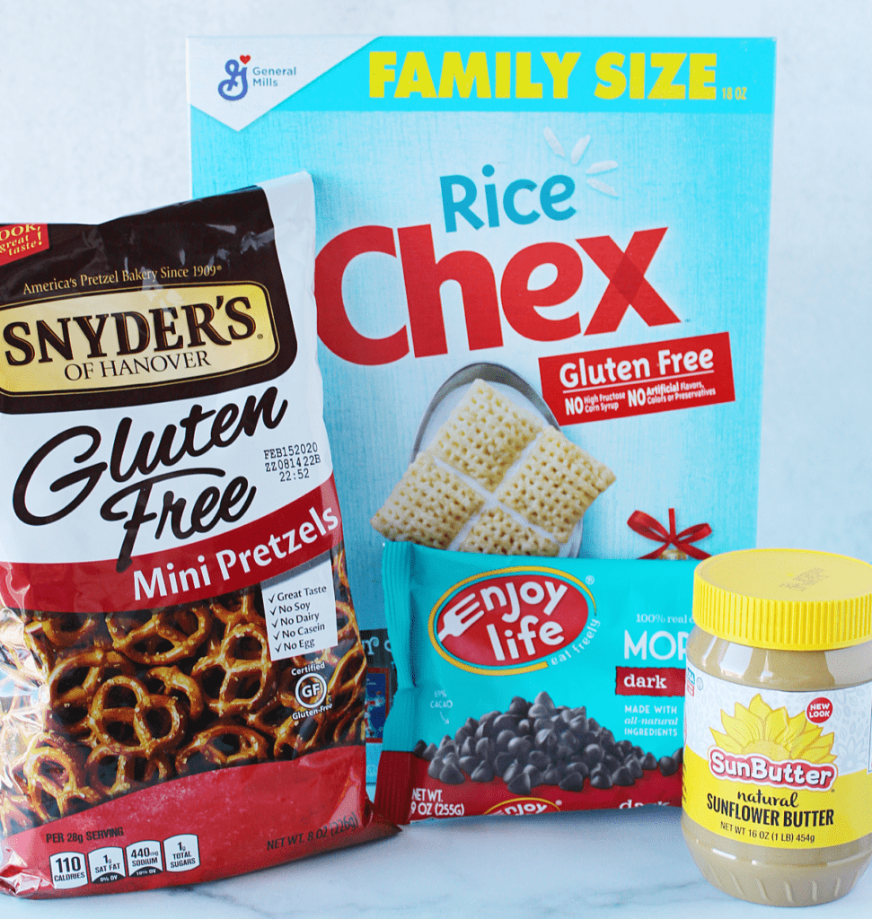 Bag of gluten free mini pretzels, box of gluten free rice Chex cereal, a bag of dairy freee Enjoy Life dark chocolate chips, and a container of nut free Sunbutter on the counter.