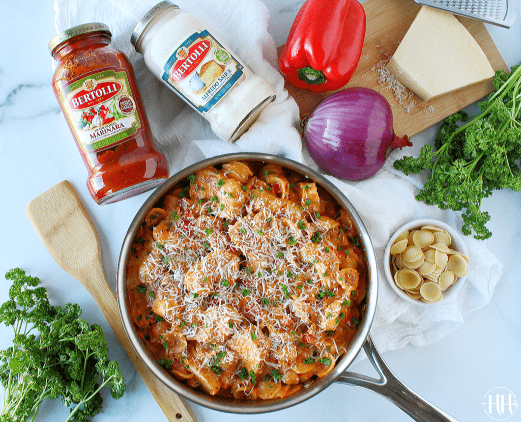 A pretty scene of a pan of finished pasta surrounded by jars of pasta sauce, vegetables, and various other ingredients made for a Valentine's Day Date Night. 