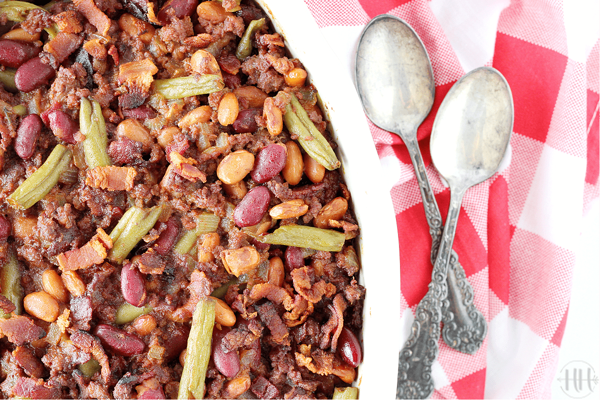 Photo of a baking dish with calico beans with spoons and a red checkered towel.