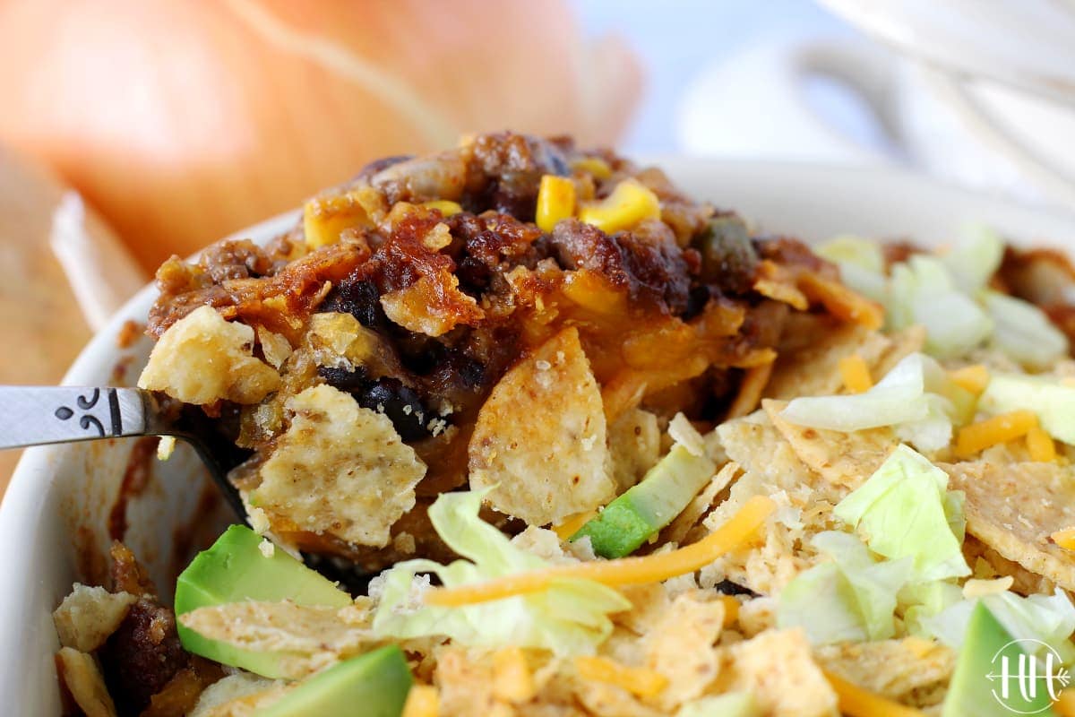 Spoonful of beef enchilada casserole topped with crushed tortilla chips, diced avocado, lettuce, and shredded cheese.