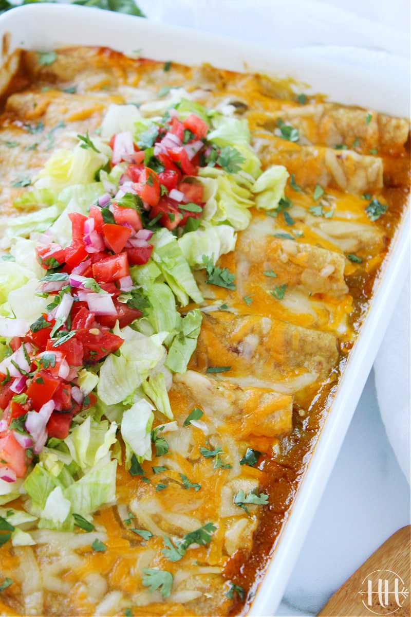 White pan full of beef enchiladas topped with lettuce and pico de gallo.