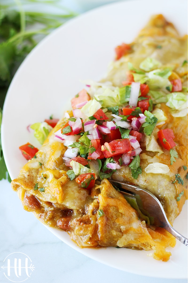 A fork cutting through a beef enchilada in green sauce topped with chopped tomatoes.