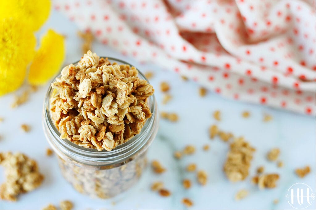 Small mason jar of vegan granola on a white countertop and polka dotted towel and flours. 