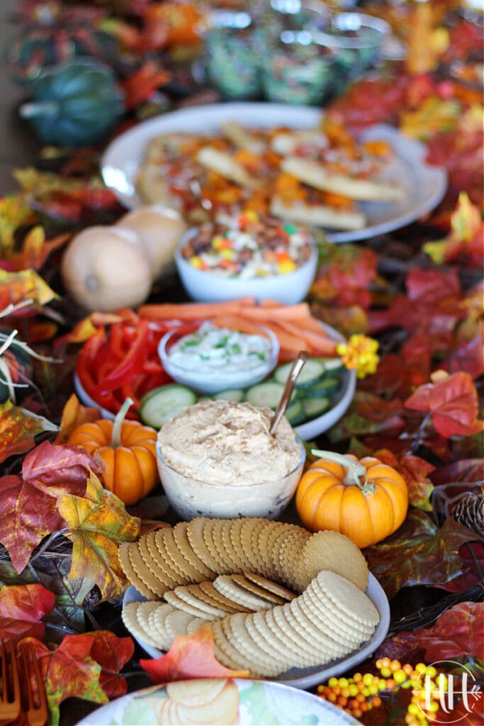 Lovely dark wood table full of friendsgiving appetizer menu ideas like pumpkin pie dip, veggies and dip, and butternut squash pizza. 