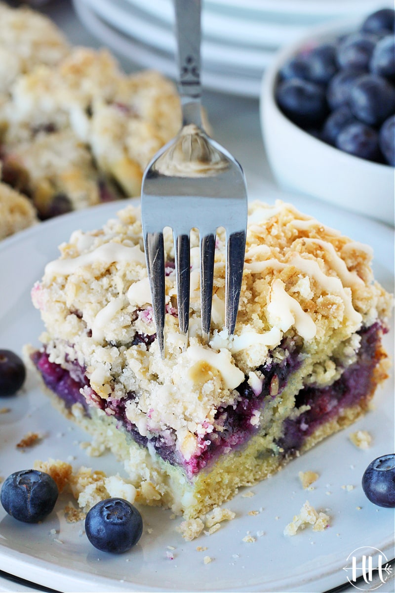 A fork slicing through a piece of blueberry coffee cake on a white plate.