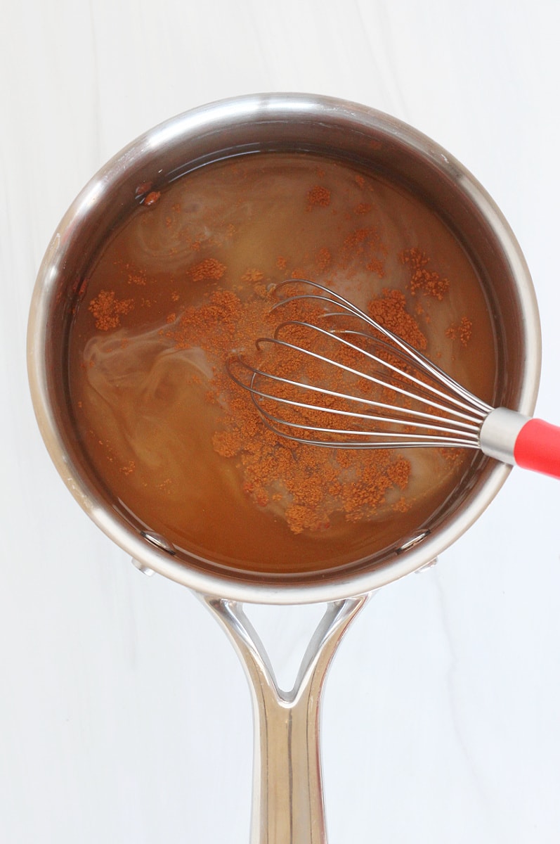 A whisk stirring cinnamon into bone broth hot cocoa ingredients in a small saucepan.