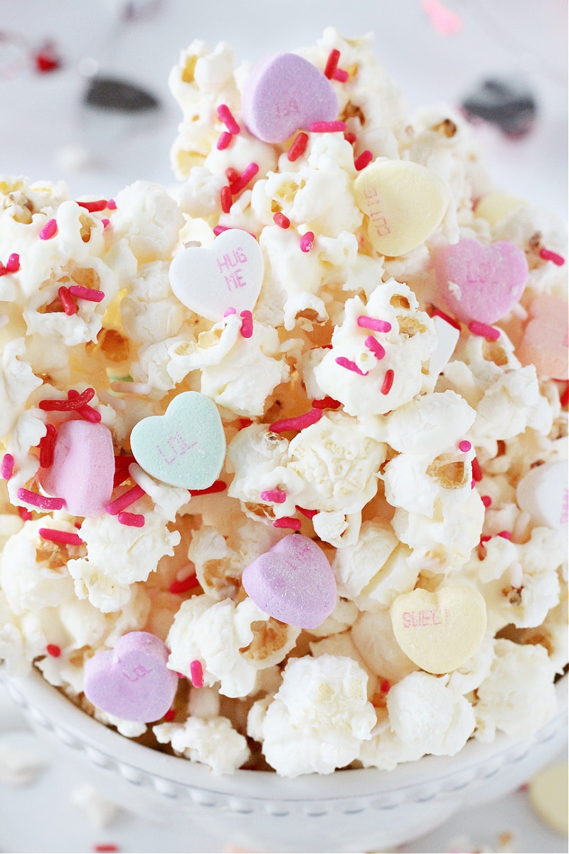 Colorful Valentine conversation hearts and white chocolate popcorn stacked in a white bowl.