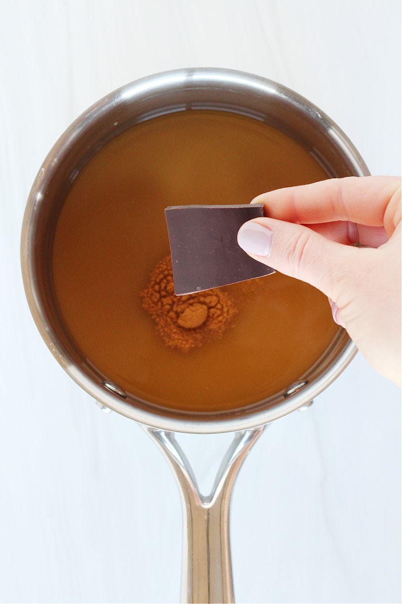 A woman's hand adding a piece of dark chocolate to a pan of bone broth.