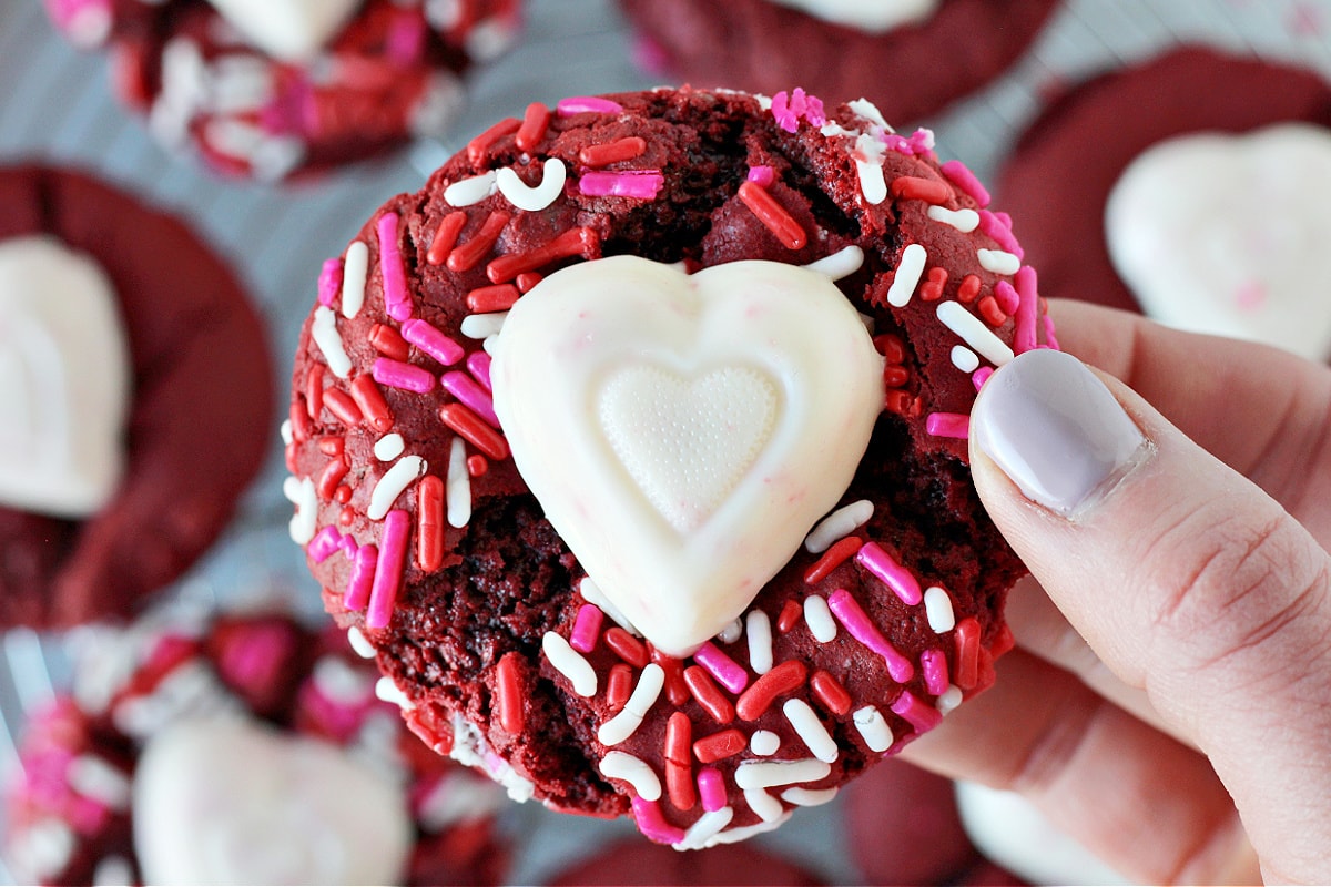 A woman's hand holding a red velvet cake mix cookie with a white chocolate heart.