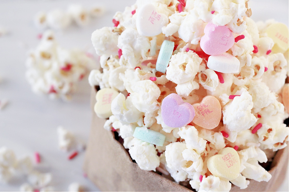White chocolate covered popcorn with conversation hearts and Valentine sprinkles in a paper bag.