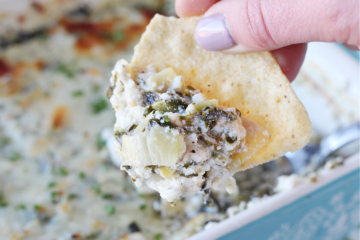 A woman's fingers holding a chip with hot spinach and artichoke dip on it.