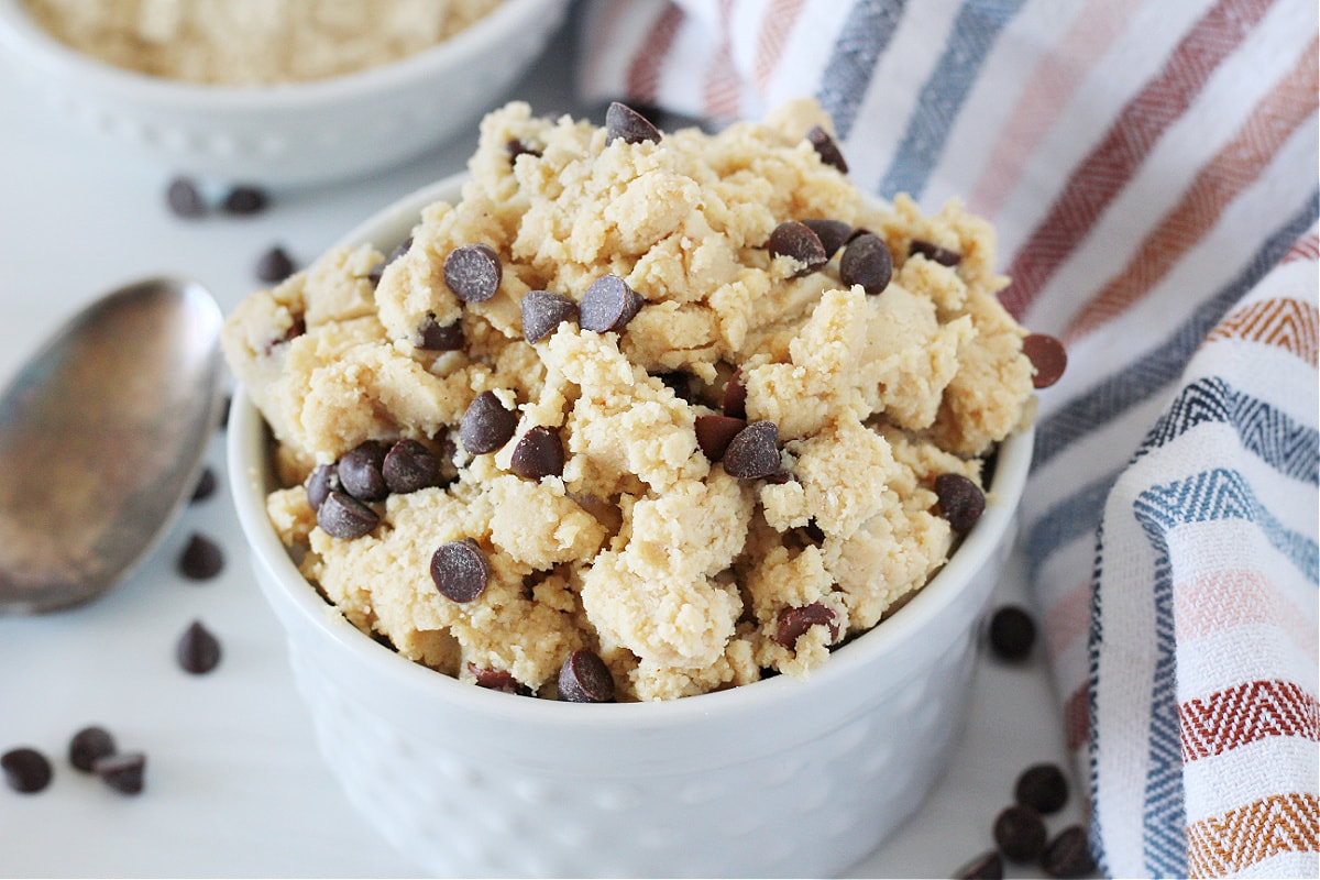 Edible chocolate chip cookie dough overflowing out of a white ceramic dish.