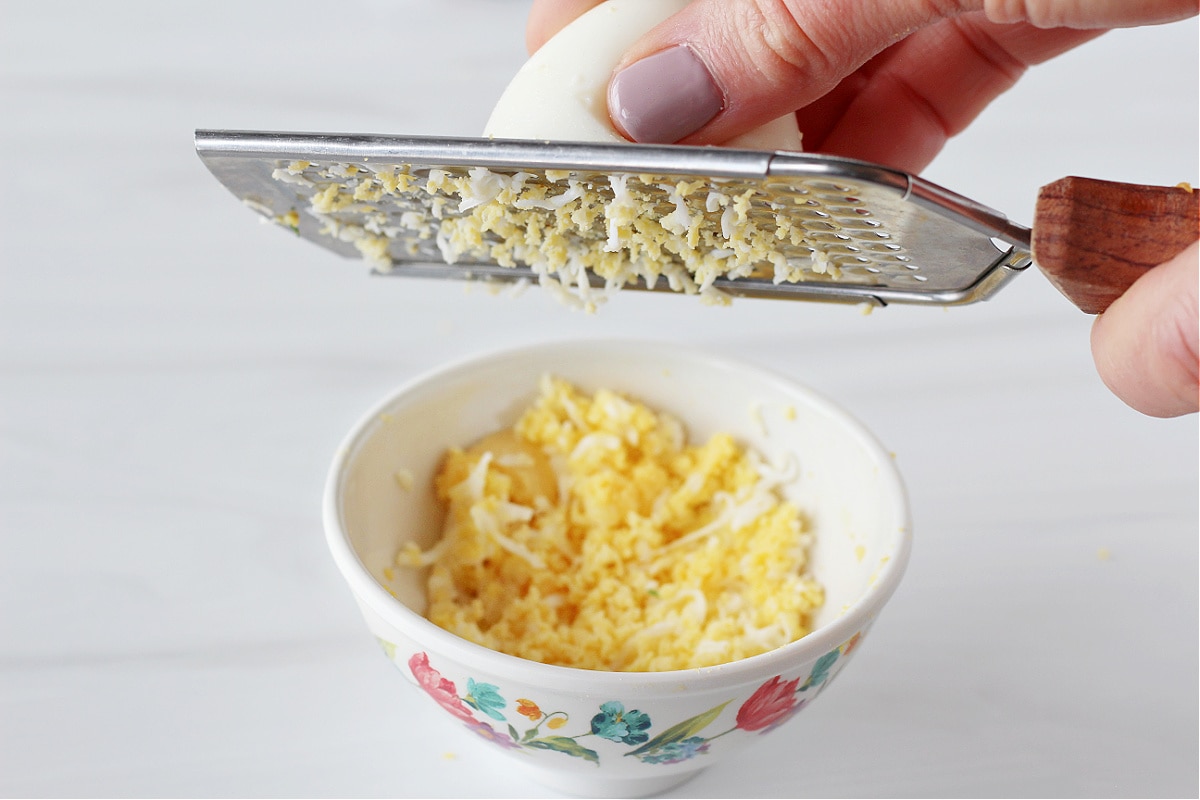 A woman's hands grating a hard boiled egg with a microplane over a small bowl.