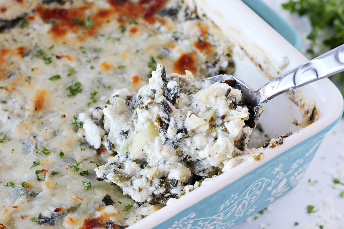 A spoon digging into a pan of baked spinach and artichoke dip in a pan.
