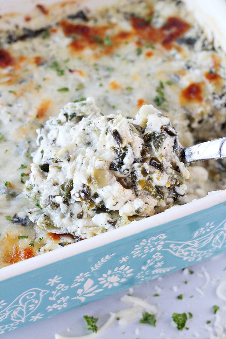 A spoon in a ceramic dish of baked creamy spinach and artichoke dip.