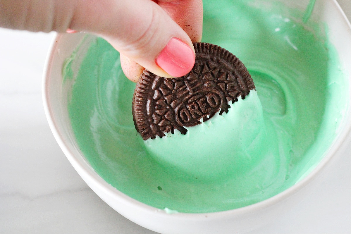 Half an Oreo being dipped in a bowl of melted green chocolate.