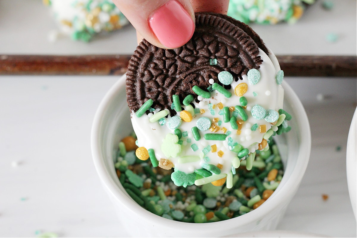 A St. Patrick's Day Oreo dipped in white chocolate being dipped in festive sprinkles.