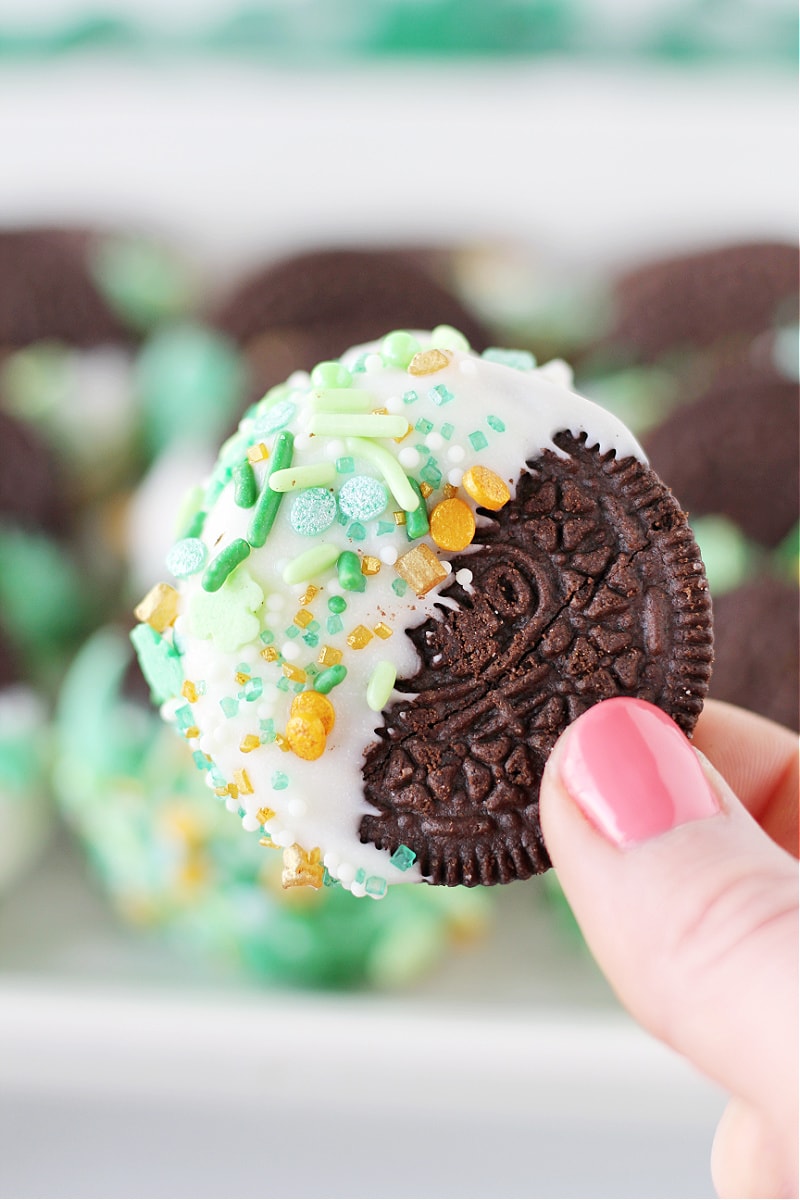A woman's fingers holding a St. Patrick's Day White Chocolate Dipped Oreo with festive sprinkles.